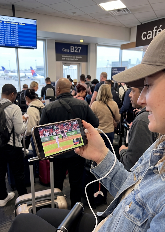 Person watching a sports game on a smartphone in an airport with other travelers around.